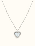 A silver heart pendant necklace featuring a milky white mother-of-pearl heart inlay, surrounded by a halo of small clear crystals on a delicate silver chain, set against a solid off-white background.