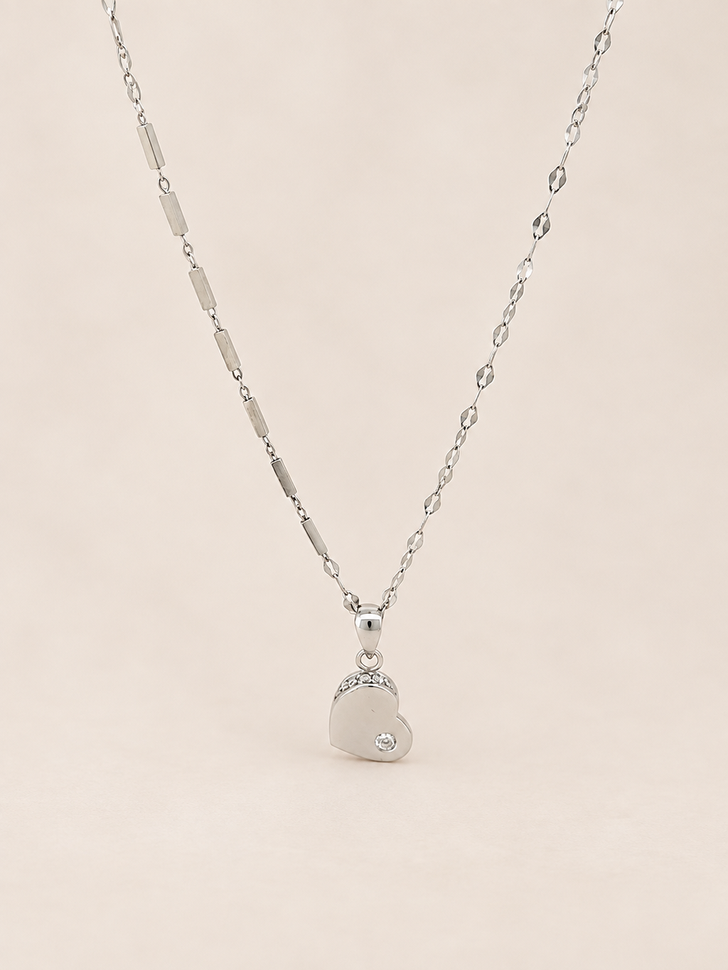 A silver heart pendant necklace with a small clear gemstone, hanging from a delicate silver chain against a soft beige background.