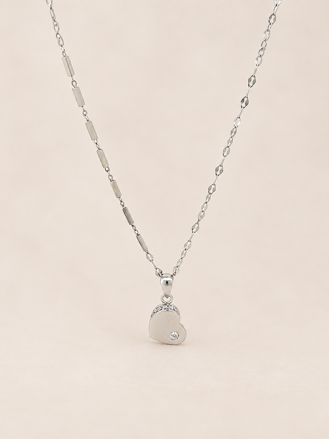 A silver heart pendant necklace with a small clear gemstone, hanging from a delicate silver chain against a soft beige background.