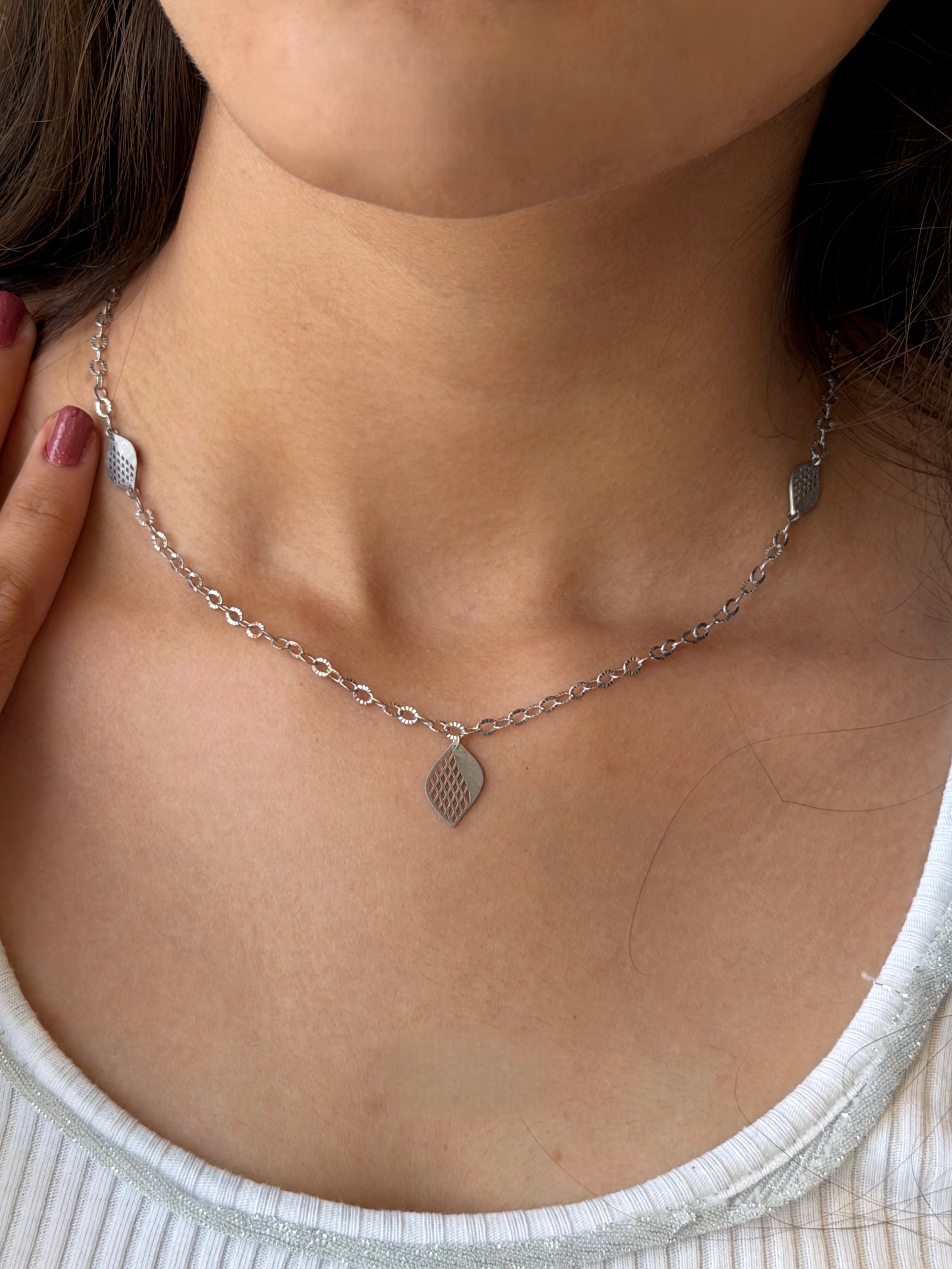 A close-up of a person's neck and upper chest wearing a delicate silver chain necklace with a small textured teardrop shaped leaf pendant.