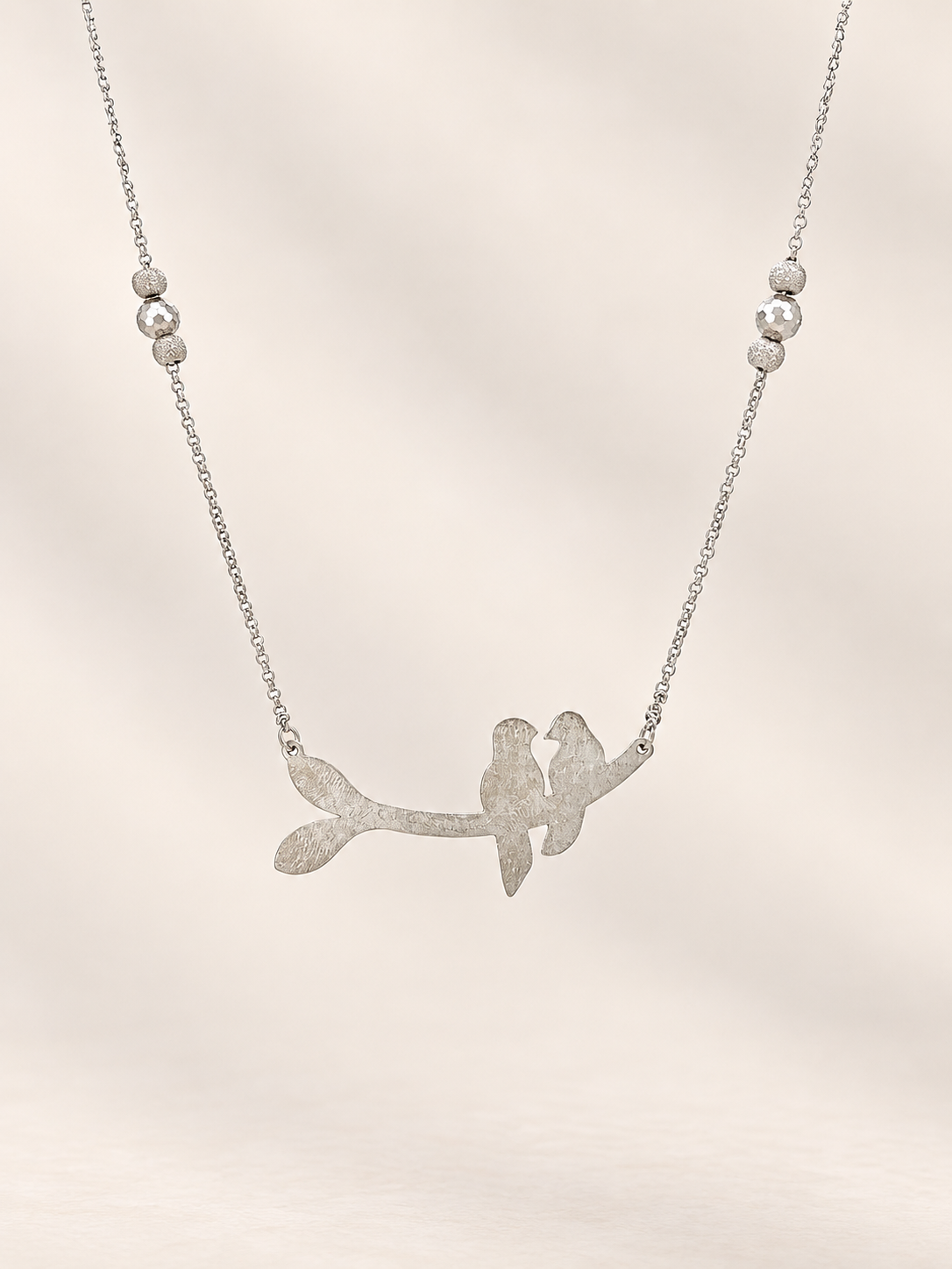 A silver necklace featuring a textured branch pendant with two small birds perched on it, suspended from a delicate silver chain with three small round beads on each side, set against a soft beige background.