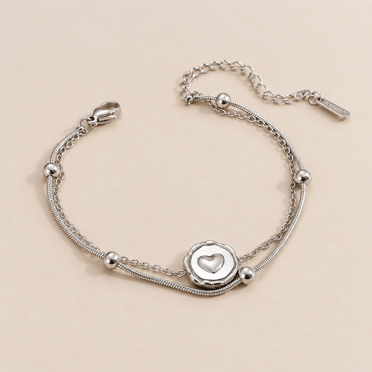 A silver double-strand chain bracelet featuring a circular heart-motif centre charm and polished silver bead details, displayed on a clean pink background.
