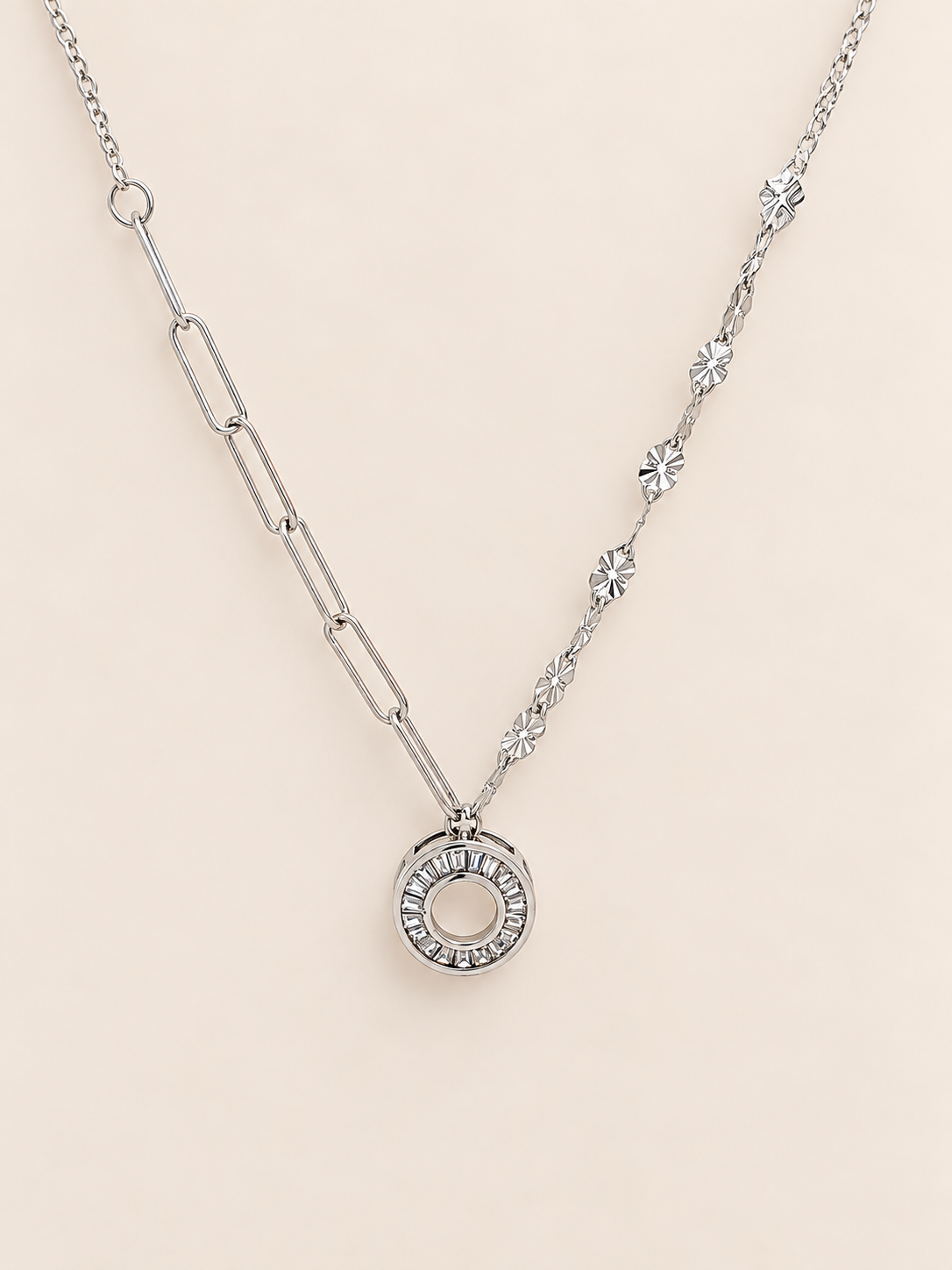 A silver-toned necklace featuring a paperclip chain segment on one side, a delicate floral star chain on the other, and a circular ring pendant encrusted with small clear stones, set against a solid pale pink background.