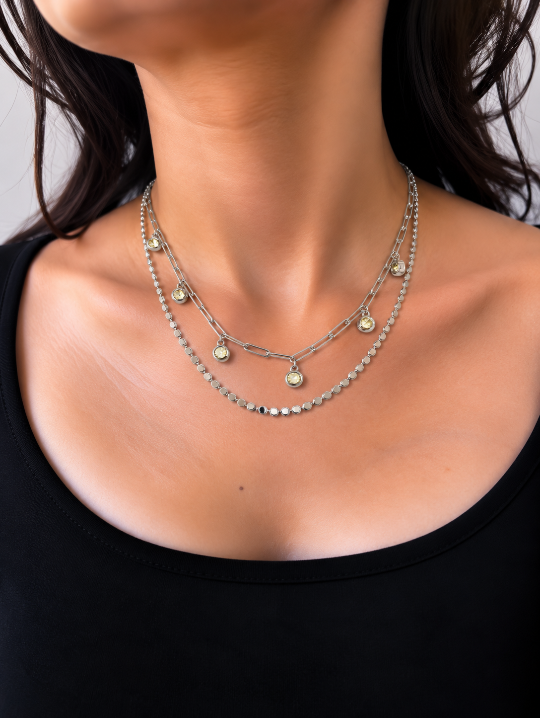 A layered silver necklace set featuring a paperclip chain, a beaded chain, and a delicate chain with small round yellow gemstone charms, worn by a model.