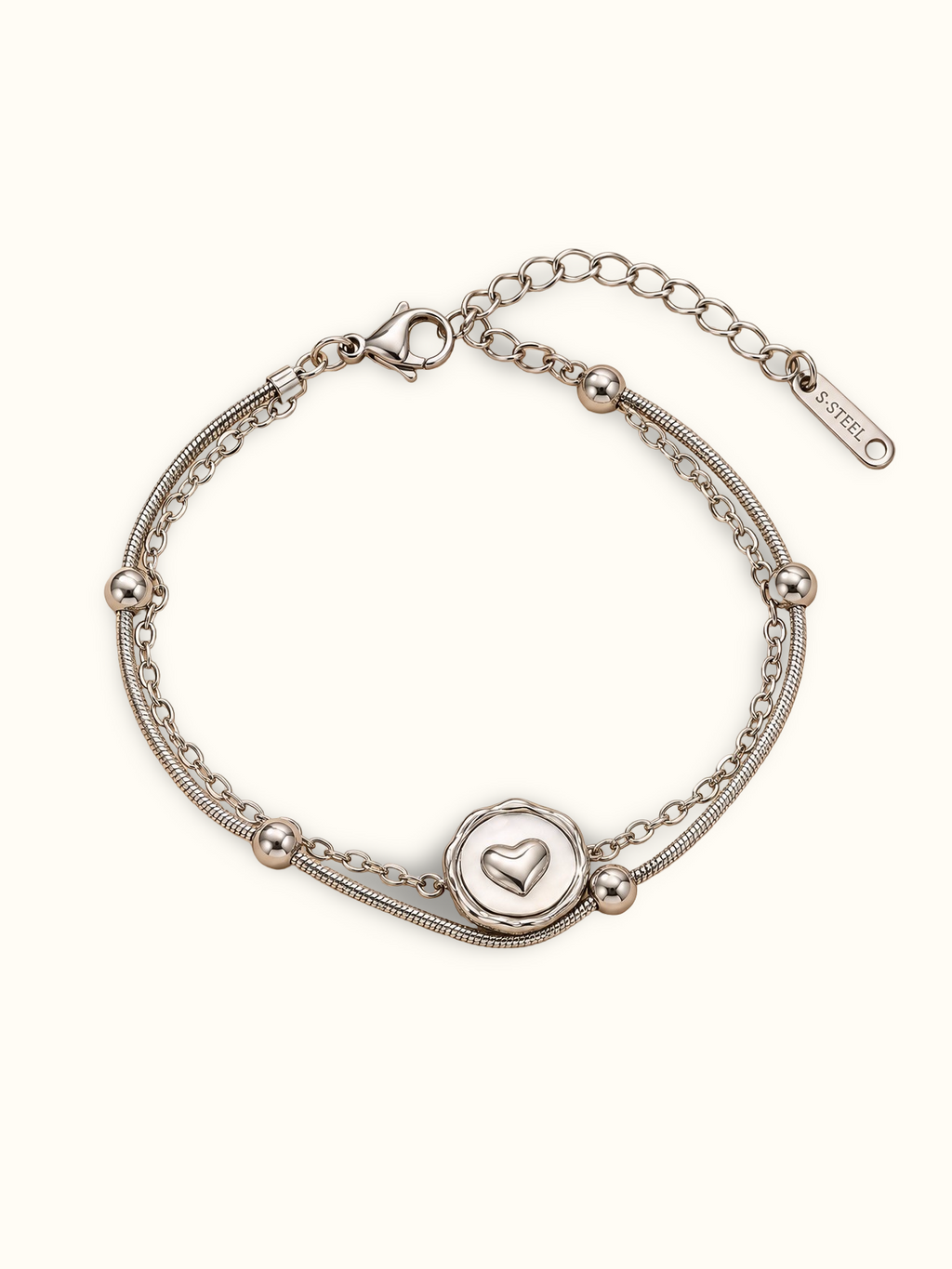 A silver double-strand chain bracelet featuring a circular heart-motif centre charm and polished silver bead details, displayed on a clean off-white background.
