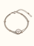 A silver double-strand chain bracelet featuring a circular heart-motif centre charm and polished silver bead details, displayed on a clean off-white background.
