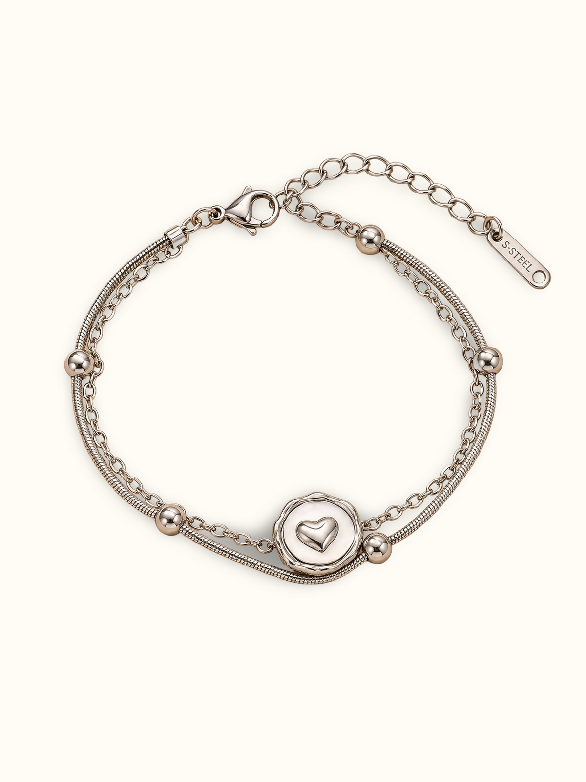 A silver double-strand chain bracelet featuring a circular heart-motif centre charm and polished silver bead details, displayed on a clean off-white background.
