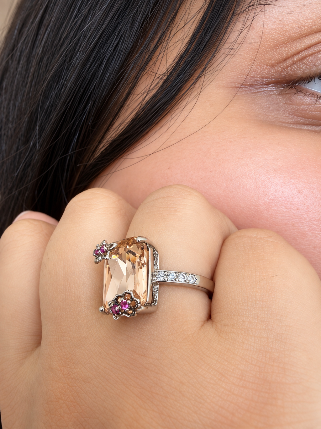 A close-up lifestyle shot of a silver ring with a large peach-colored rectangular gemstone and small pink gemstones, worn on a models finger.
