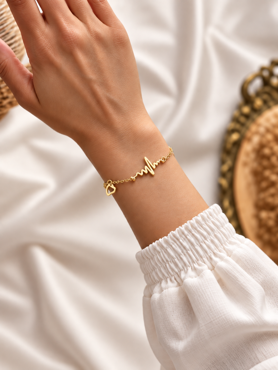 A delicate gold chain bracelet with a heartbeat line and heart charm, worn on a woman's wrist. The person is wearing a white long-sleeve top, and the background consists of draped white fabric with a decorative woven basket visible on the sides.