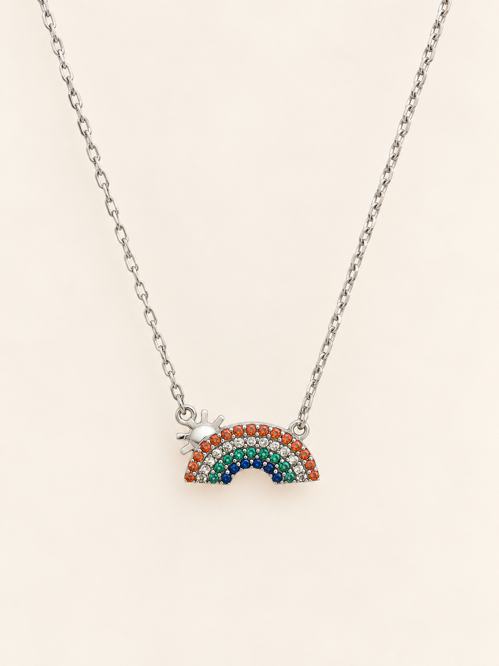 A silver chain necklace featuring a small rainbow charm paved with multicolored rhinestones and a tiny silver star accent, displayed on a light cream background.