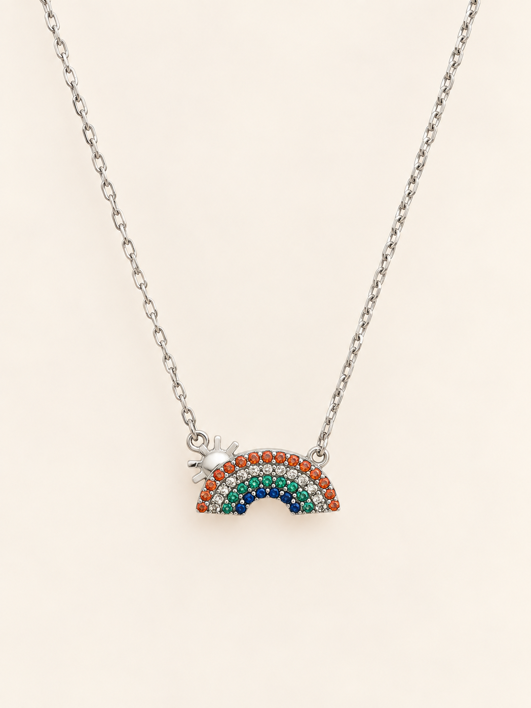 A silver chain necklace featuring a small rainbow charm paved with multicolored rhinestones and a tiny silver star accent, displayed on a light cream background.