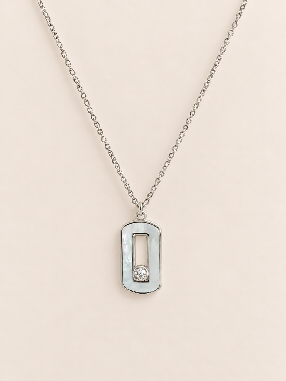 A silver-toned necklace with a rectangular pendant featuring a vertical cutout and a small clear stone, displayed on a soft beige background.