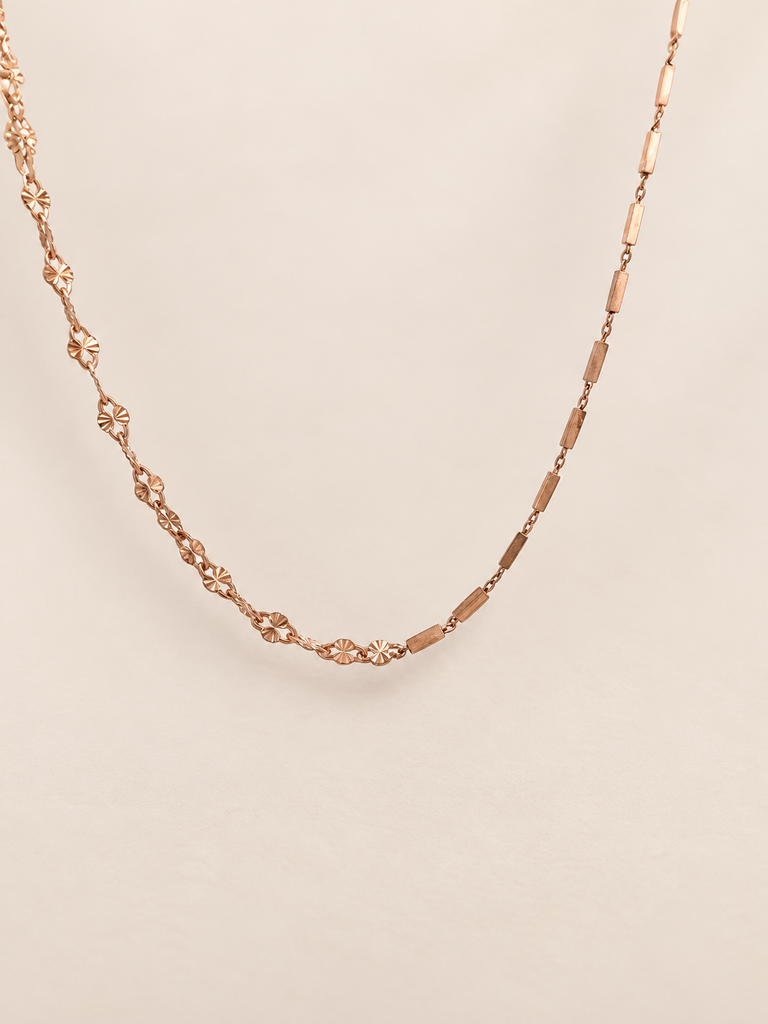 A delicate rose gold chain necklace featuring alternating textured floral links and elongated oval links, displayed against a solid light beige background.