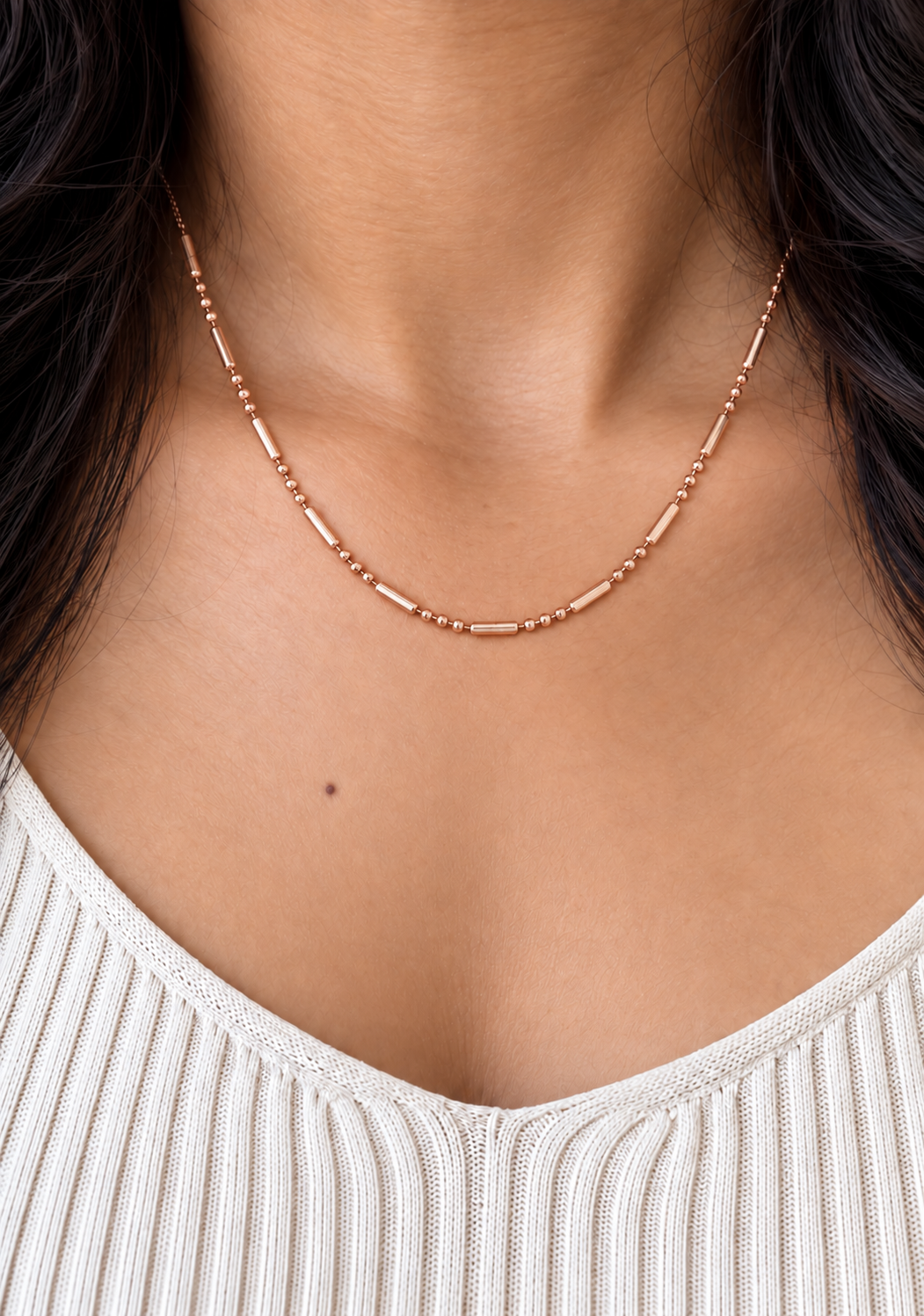 A close-up of a woman's neck wearing a delicate rose gold beaded chain necklace over a cream ribbed knit top.
