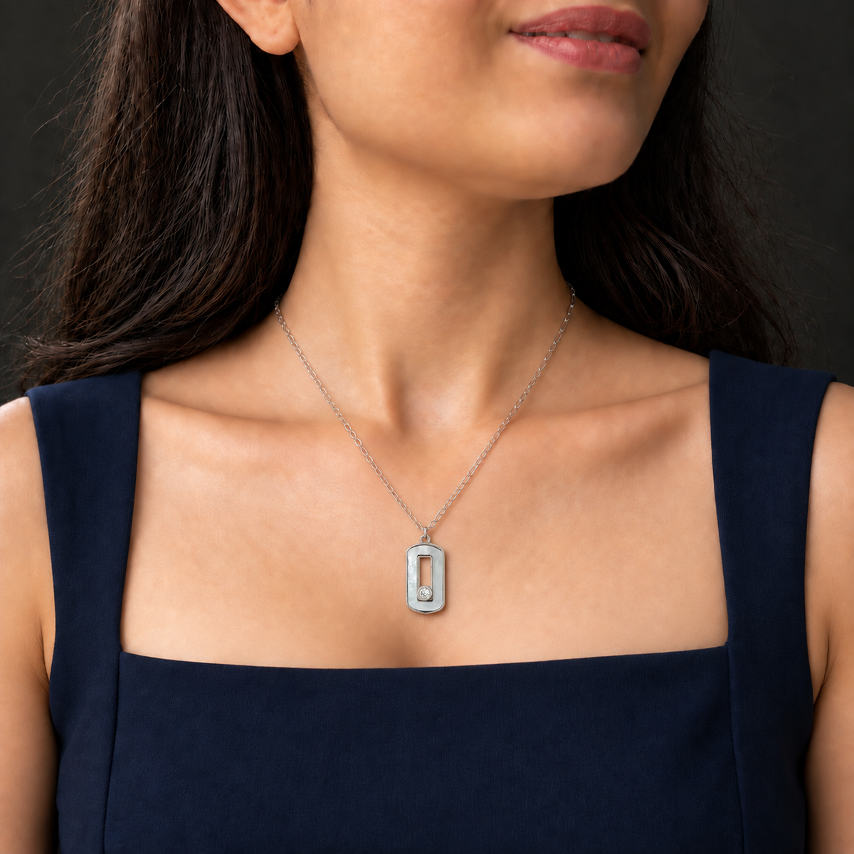 A close-up of a woman wearing a silver-toned rectangular pendant necklace with a central cutout, styled with a navy blue sleeveless top against a dark background.
