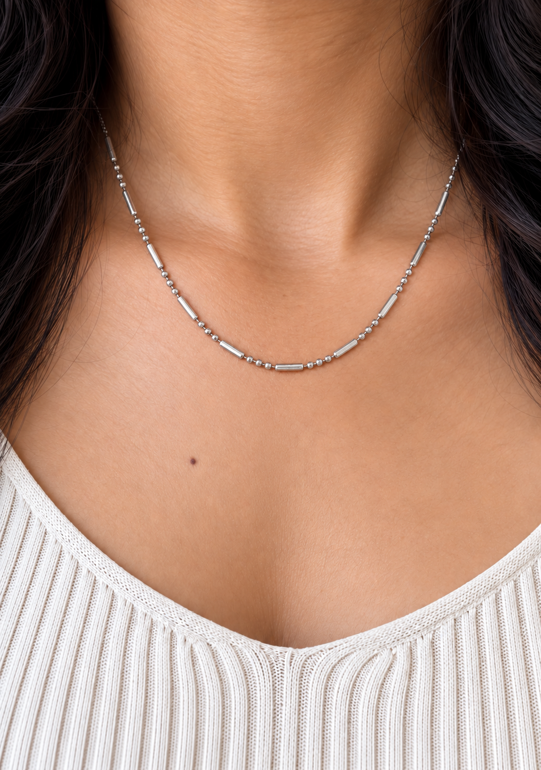 A close-up of a model's neck wearing a delicate silver chain necklace with small cylindrical links, paired with a white ribbed knit top.
