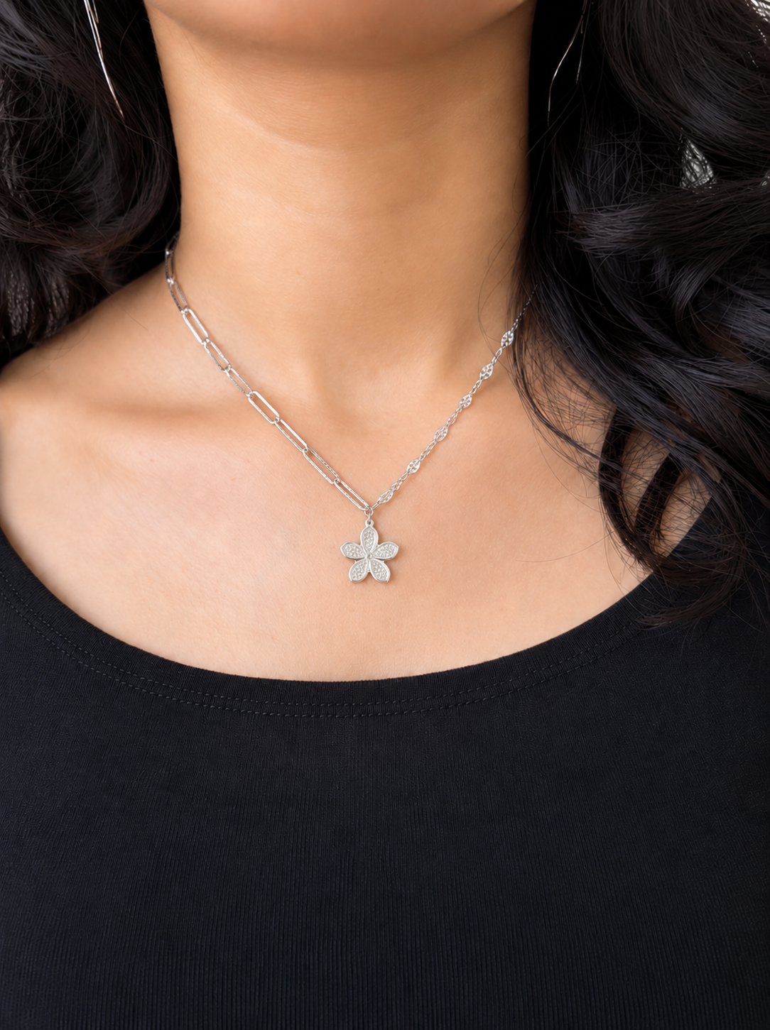 A close-up of a model wearing a silver chain necklace with a small flower-shaped pendant, paired with a black top.