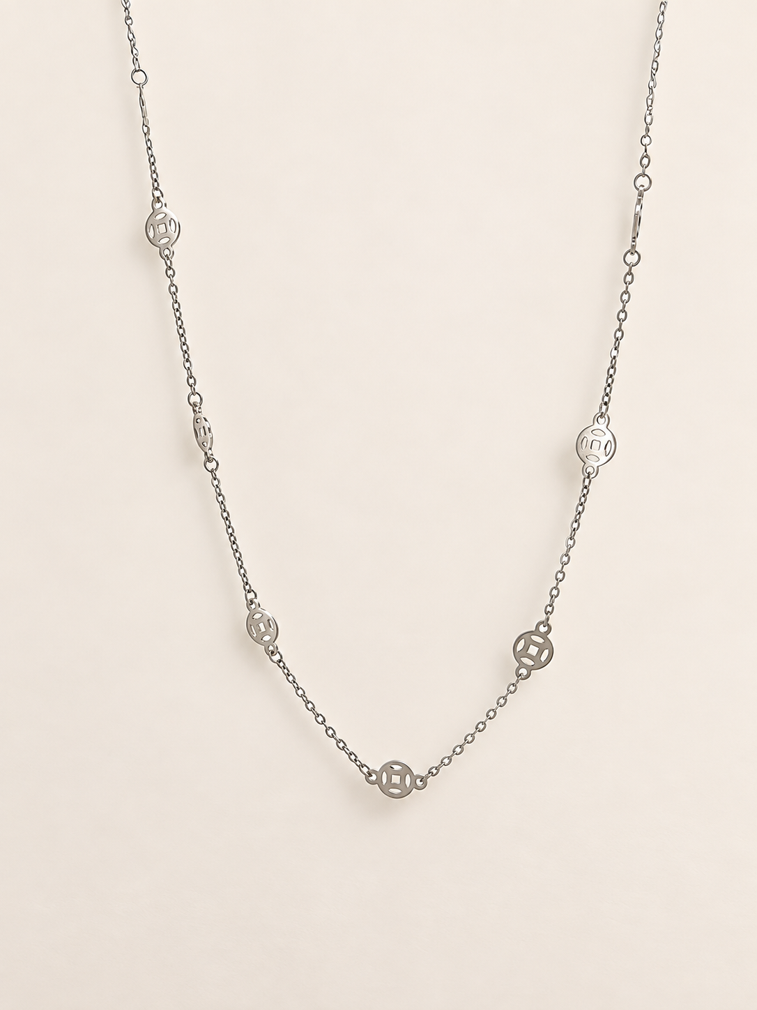 A delicate silver chain necklace featuring multiple small circular medallion charms, displayed against a solid, warm off-white background.
