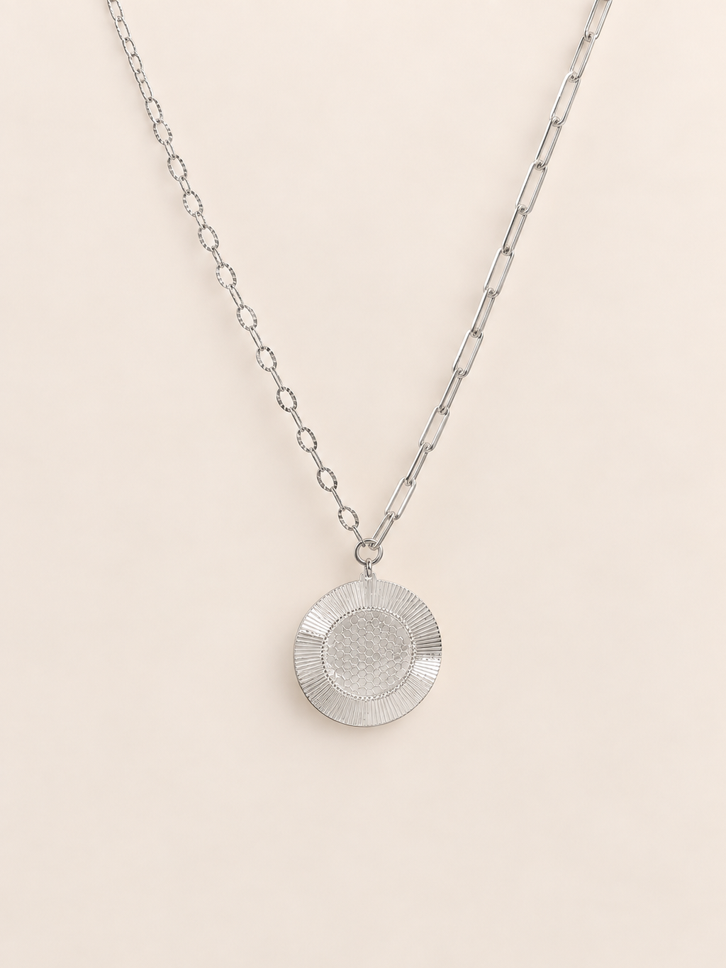A silver pendant necklace featuring a textured circular disc on a paperclip chain, displayed against a solid pale pink background.