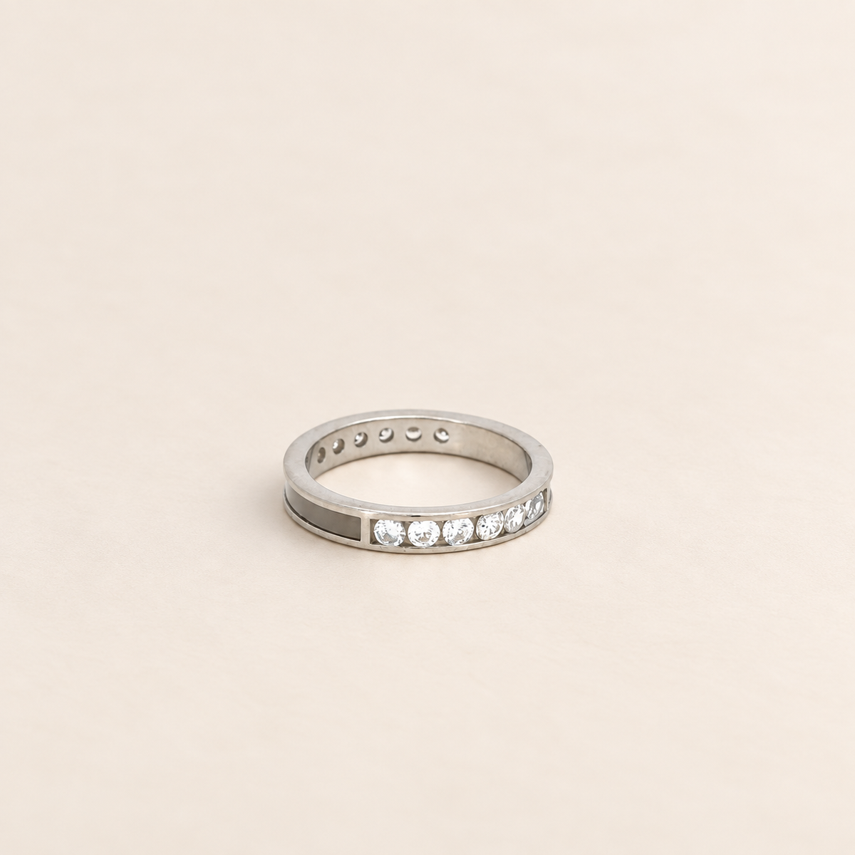 A silver band ring featuring a row of clear round-cut gemstones on one side and a row of small round inset stones on the other, set against a solid light beige background.
