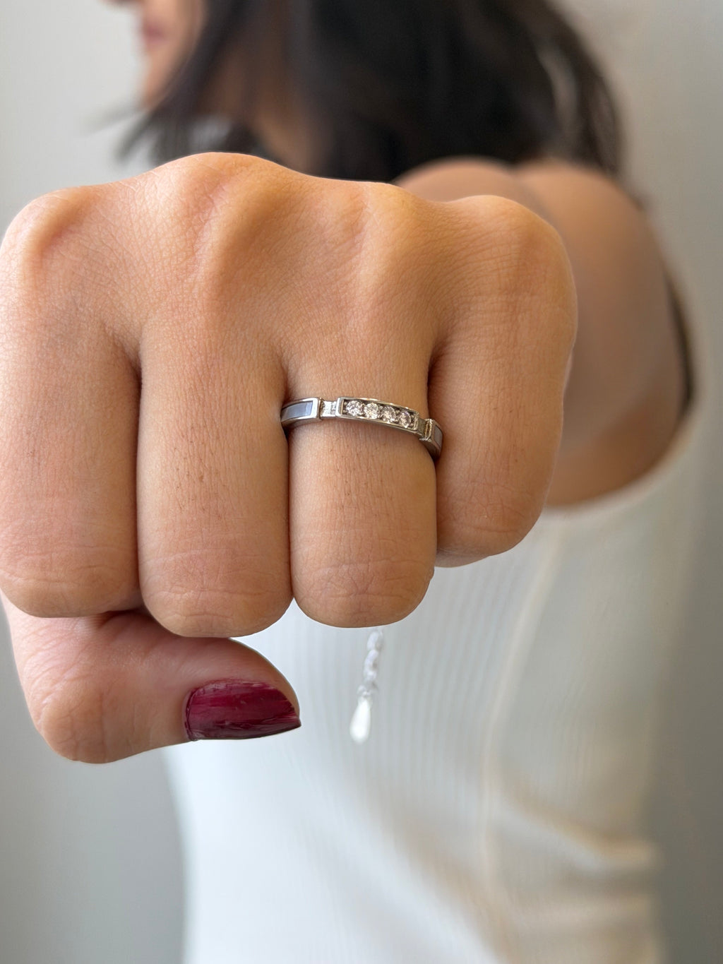 A close-up of a person's hand wearing a silver band ring with a row of small clear stones, against a soft light gray background.
