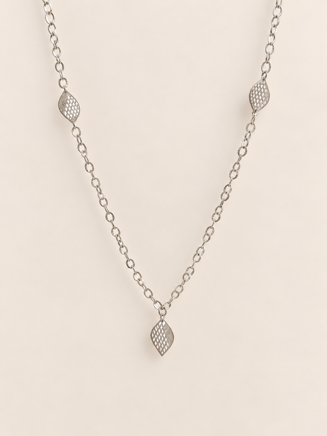 A delicate silver chain necklace featuring leaf patterned teardrop-shaped charms spaced along the chain, displayed against a solid, warm off-white background.