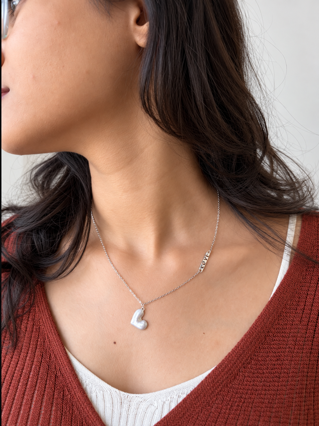 A close-up lifestyle shot of a woman wearing a delicate silver necklace with a small silver heart-shaped pendant and a small silver text charm that reads 'LOVE'.