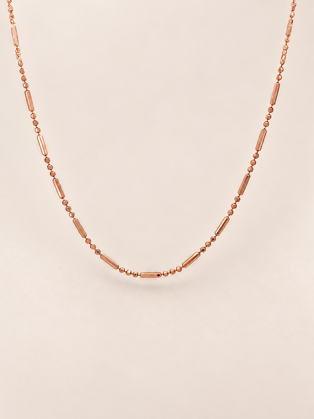 A delicate rose gold necklace featuring alternating small round beads and elongated barrel-shaped beads, displayed against a solid light beige background.