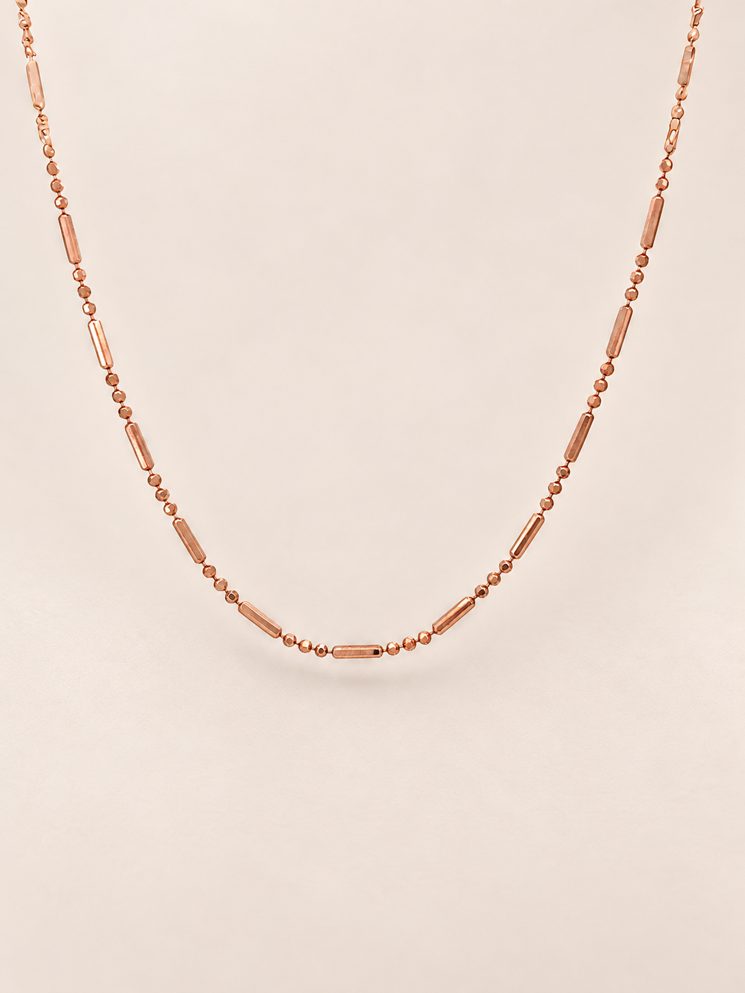 A delicate rose gold necklace featuring alternating small round beads and elongated barrel-shaped beads, displayed against a solid light beige background.