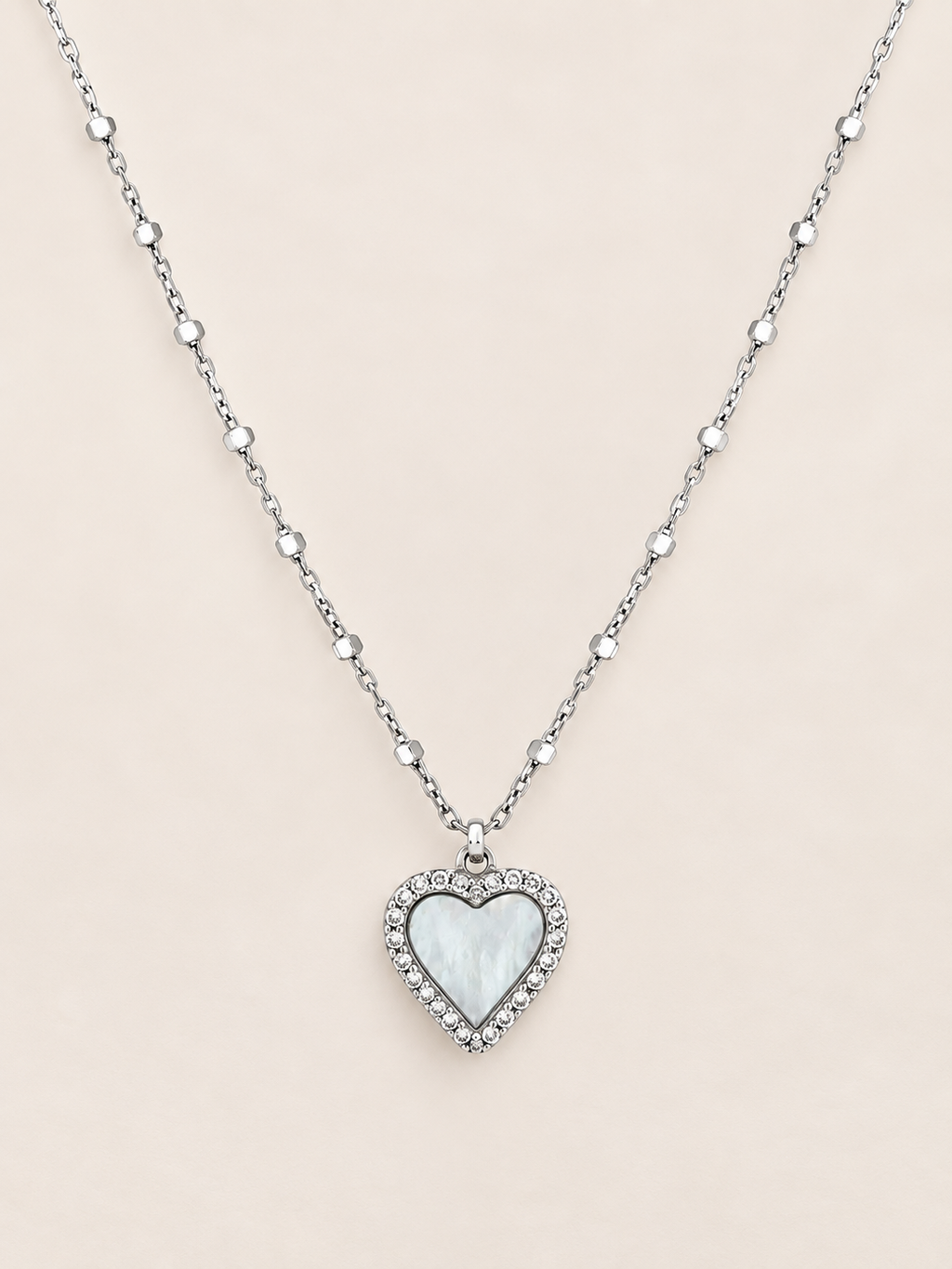 A silver heart pendant necklace featuring a white mother-of-pearl center surrounded by small clear crystals, suspended on a delicate silver beaded chain against a light beige background.