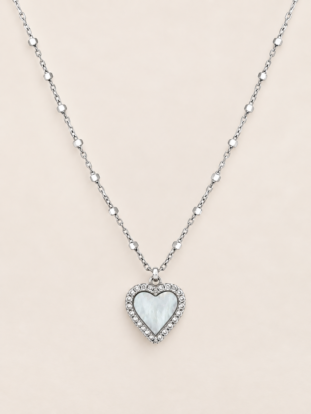 A silver heart pendant necklace featuring a white mother-of-pearl center surrounded by small clear crystals, suspended on a delicate silver beaded chain against a light beige background.