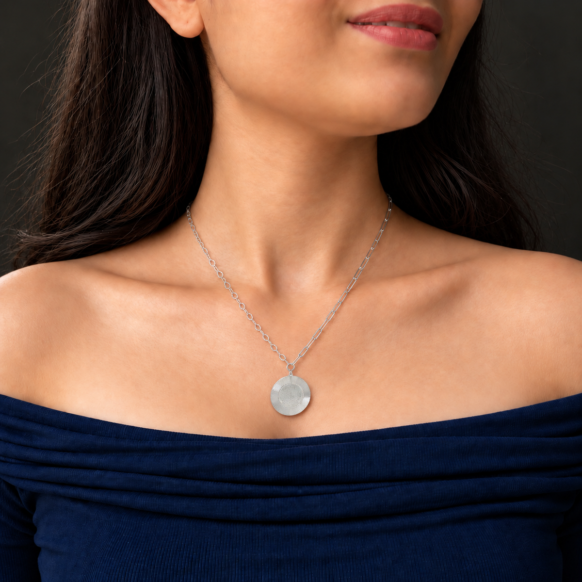 A silver pendant necklace featuring a textured circular disc on a paperclip chain, worn by a model