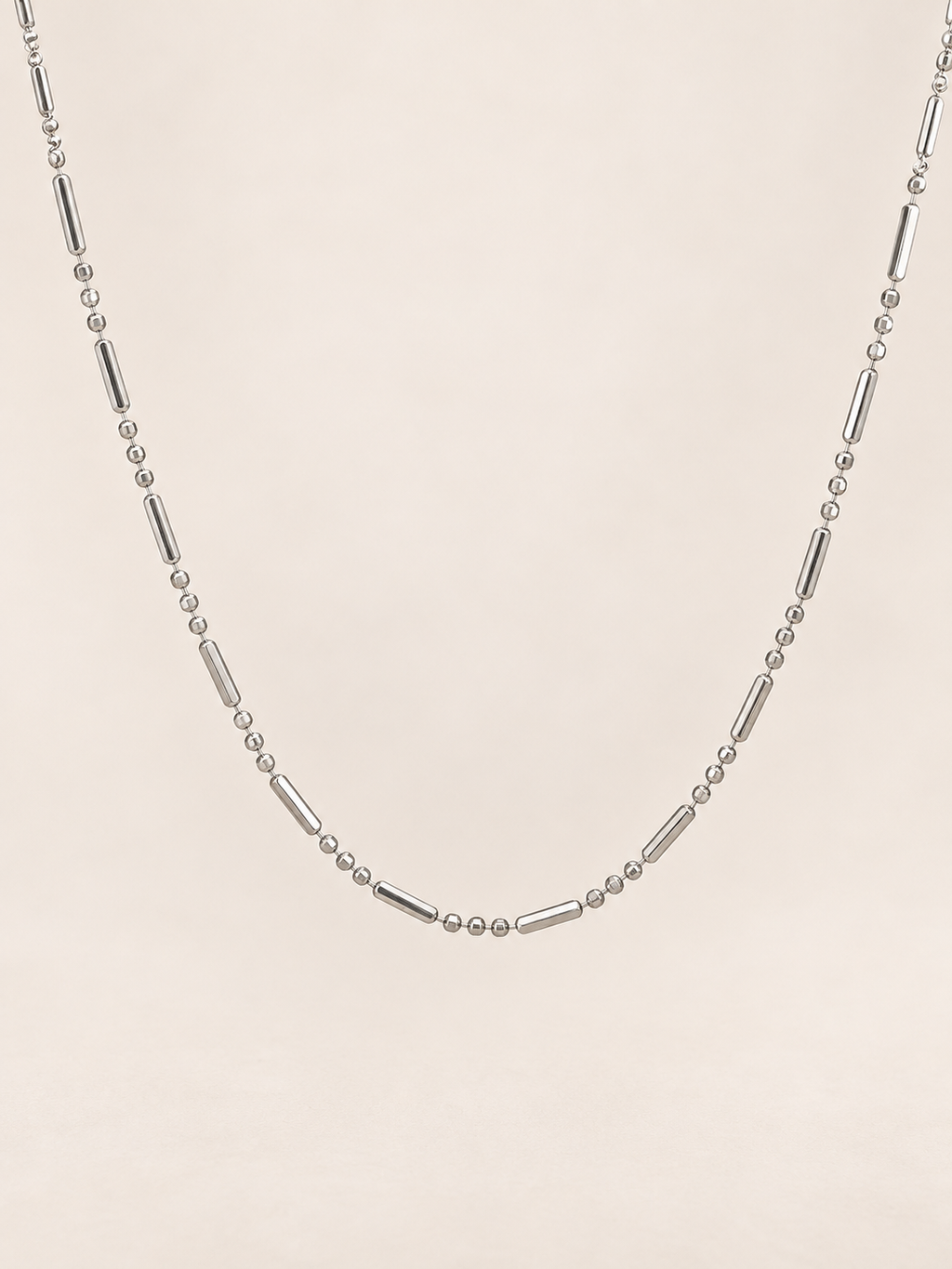 A silver-toned beaded necklace featuring alternating small round beads and elongated tube beads, displayed against a solid, warm off-white background.