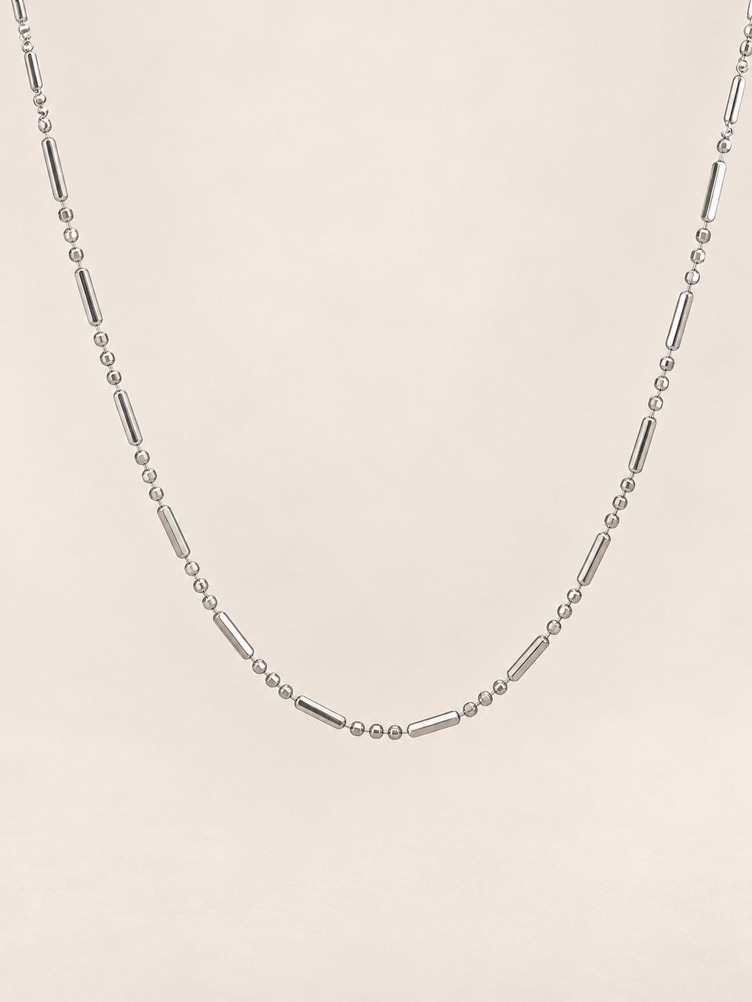 A silver-toned beaded necklace featuring alternating small round beads and elongated tube beads, displayed against a solid, warm off-white background.
