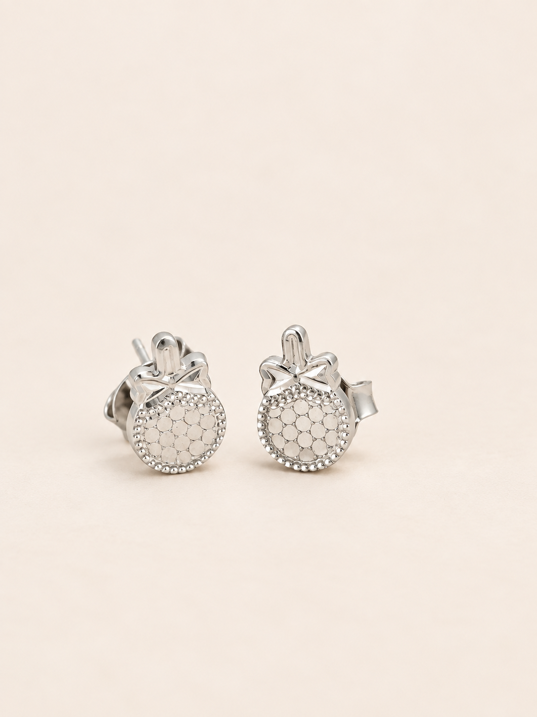 A pair of silver stud earrings with round pavé-set clear stones and a small bow detail on top, displayed on a light beige background.