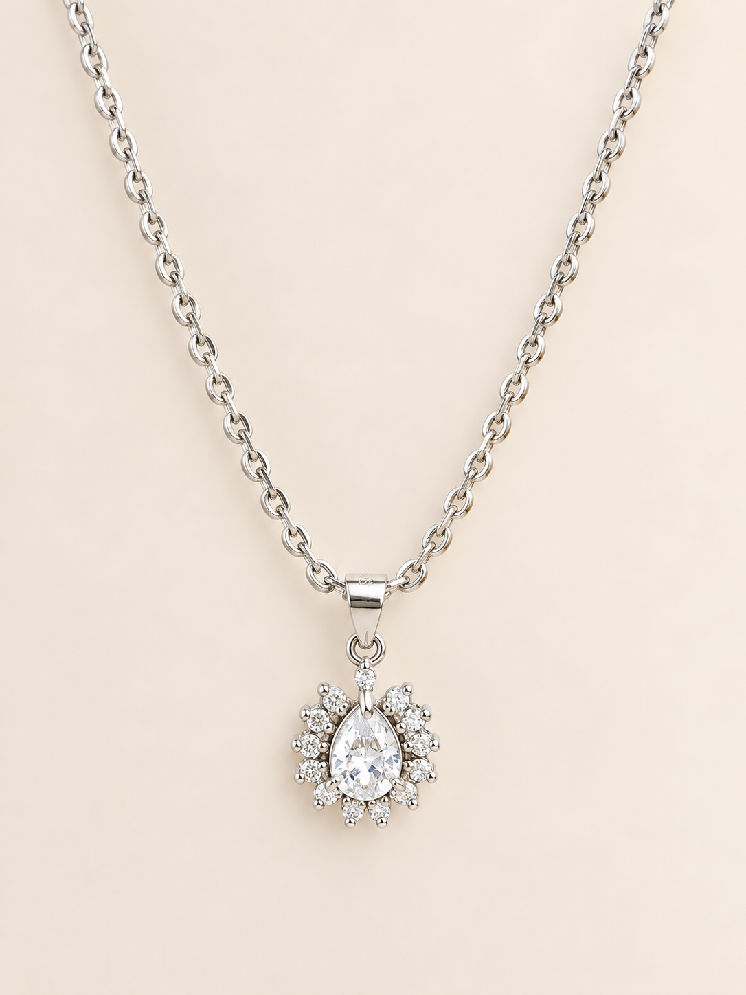 A silver-tone chain necklace with a teardrop-shaped clear gemstone pendant surrounded by a halo of small round stones, set against a solid pale pink background.