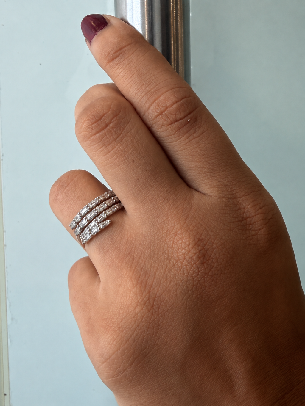 A close-up of a hand wearing three slim silver bands rings set with small clear stones, holding a metallic handle against a pale blue background.