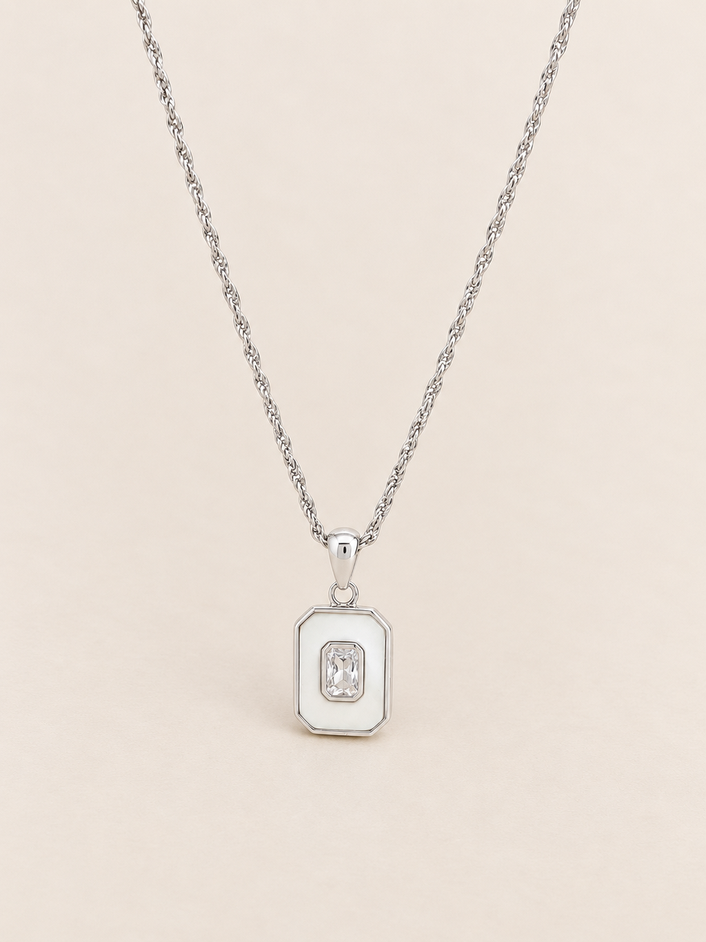 A silver pendant necklace featuring a small rectangular charm with a central oval inset, displayed on a delicate silver chain against a pink background.
