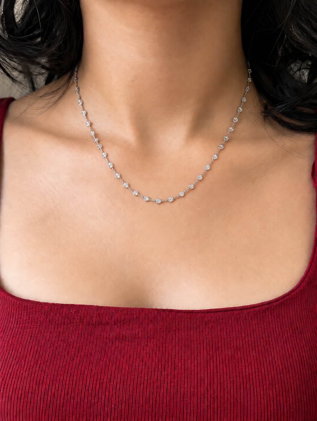 A delicate silver tennis necklace with small round-cut stones, shown worn on a model's neck over a red ribbed top.