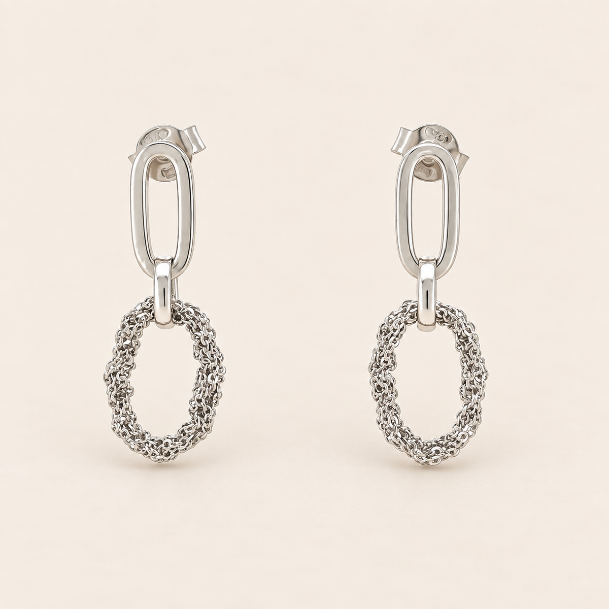 A pair of silver drop earrings with a polished oval post and a textured, openwork oval dangle, shown on a light beige background.